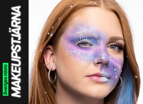 En person med galaxinspirerad makeup i lila, blå och silvertoner med vita fransar, små glittrande stenar och stjärnliknande detaljer över ansiktet och i håret.
