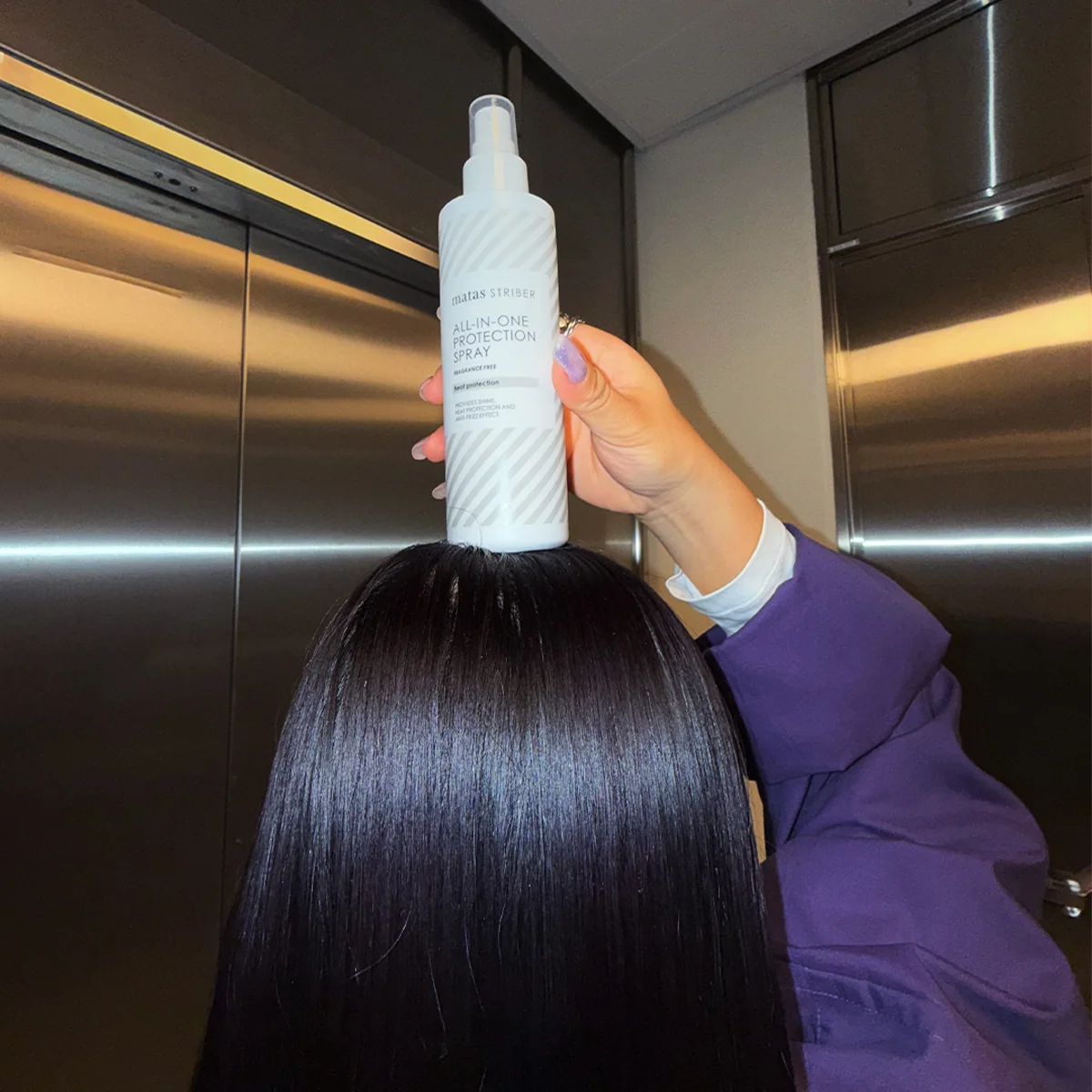 Käsi pitelee Matas Striber All-In-One Protection Spray -pulloa kiiltävän, tumman ja sileän hiuksen yläpuolella hississä.