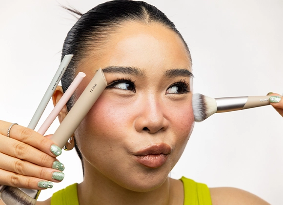 Person med mørkt hår satt opp i lav knute påfører highlighter med sminkekost på kinnbenet. Personen holder flere beige og lyse sminkekoster i én hånd.