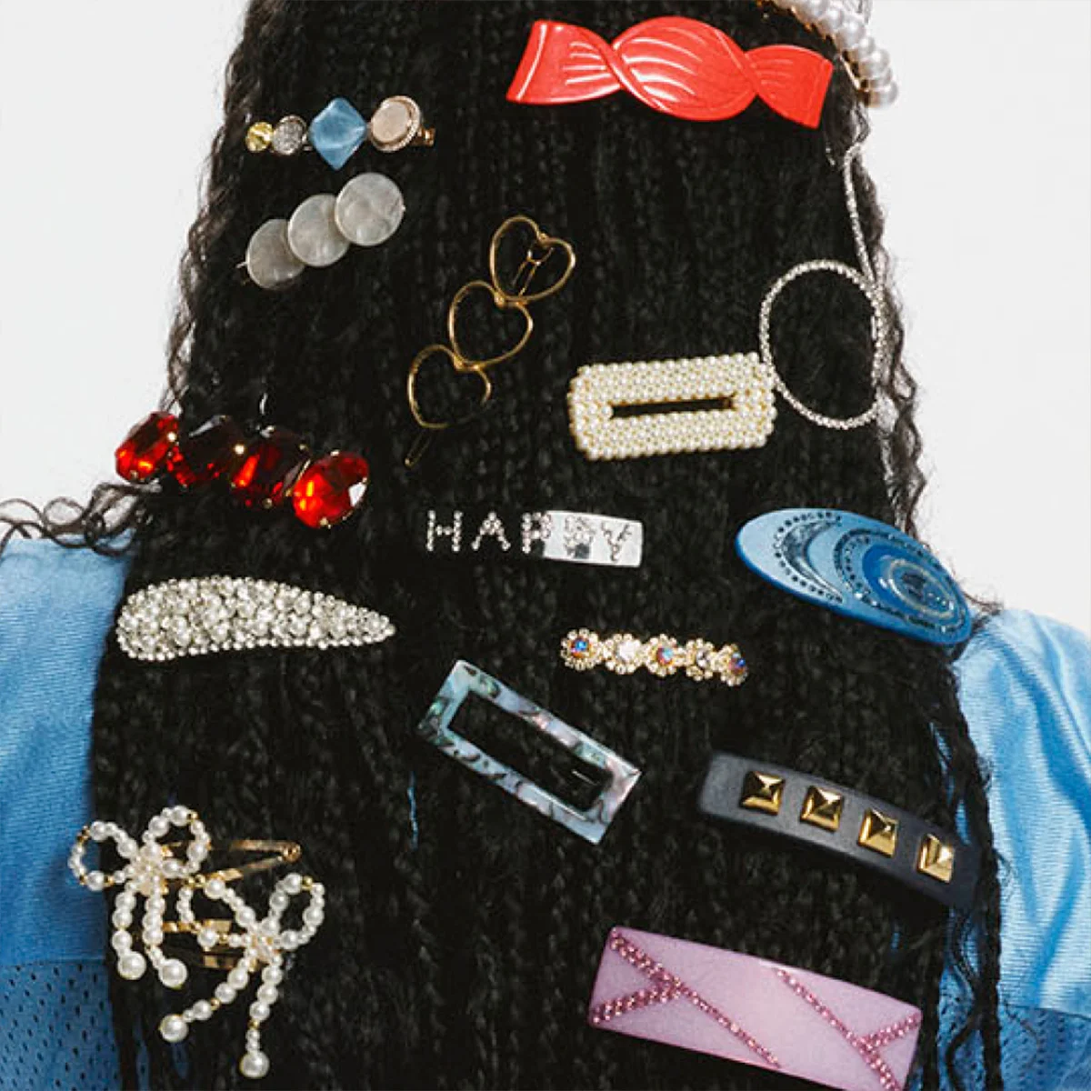 En person sedd bakifrån med långt tvinnat hår fullt dekorerat med färgstarka hårklämmor, pärlspännen, glittrande clips och formade accessoarer i olika stilar, bärandes en blå topp med meshärmar.