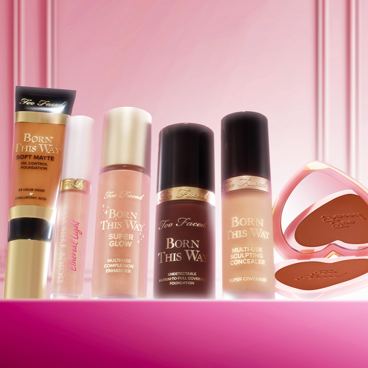 Too Faced -kasvomeikkejä rivissä vaaleanpunaisella taustalla, mukana meikkivoide, peitevoide ja aurinkopuuteri.