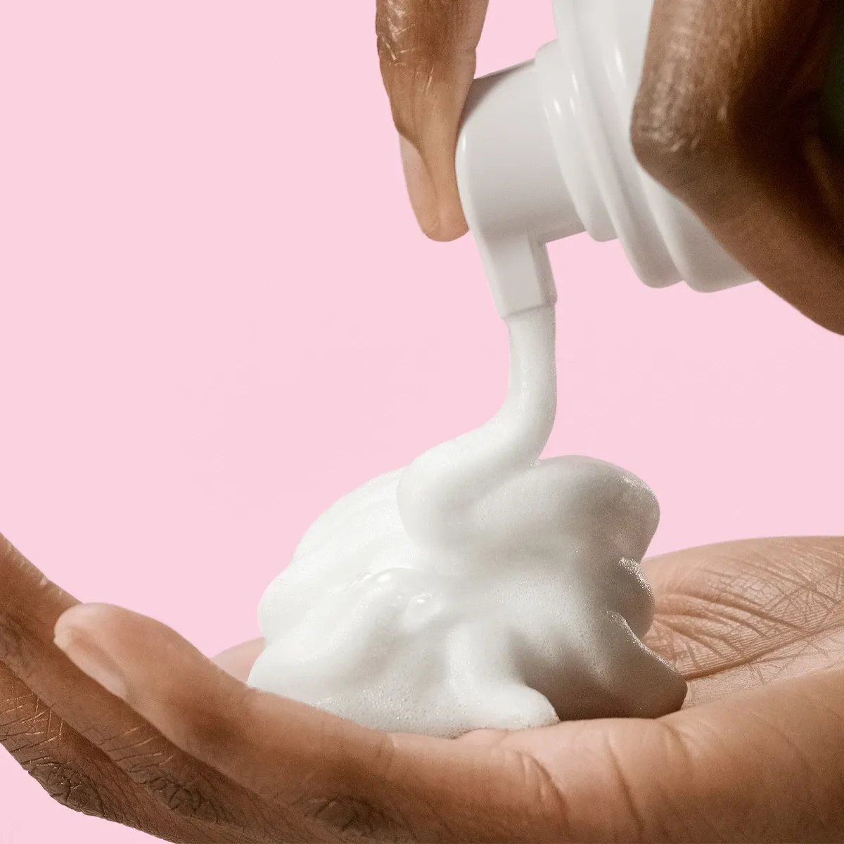 En hand som trycker ut skum från en hudvårdsprodukt till en annan hand mot en rosa bakgrund.