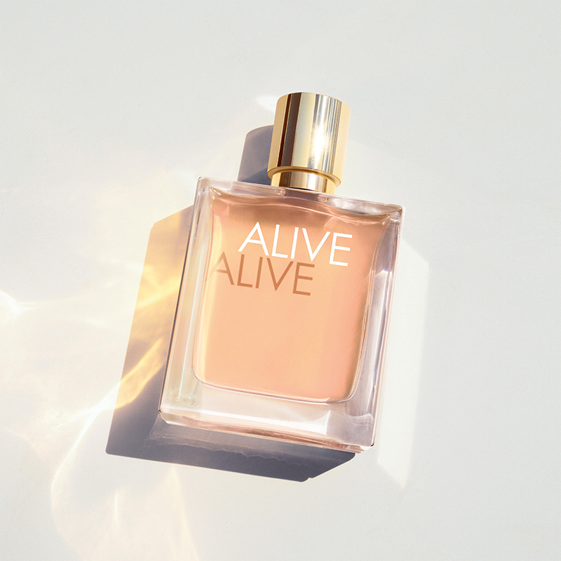 BOSS Alive Eau de Parfum läpinäkyvässä lasipullossa, jossa on kullanvärinen korkki, on vaaleaa taustaa vasten valonheijastusten ympäröimänä. Näkyvä teksti: "ALIVE" ja "ALIVE" pullossa.