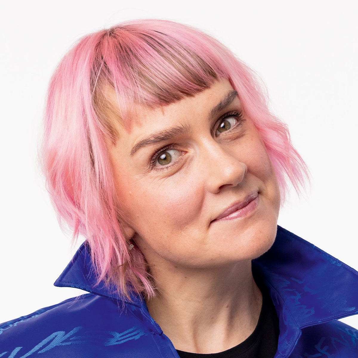 Person med kort rosa hår og lugg, fotografert mot en lys grå bakgrunn. Personen har på seg en blå jakke over en svart genser og ser mot kameraet i et skjønnhetsinspirert portrett.