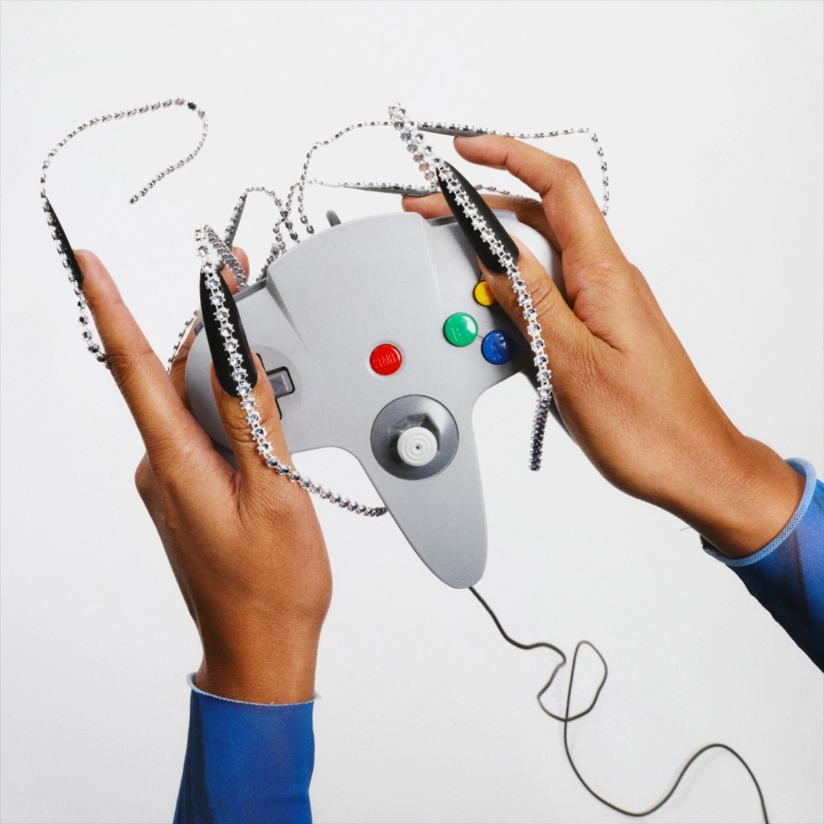 Kaksi kättä pitelee harmaata peliohjainta, jossa on värillisiä painikkeita. Käsien ja ohjaimen päällä on hopeanvärisiä ketjuja ja kimaltavia koristeita vaaleaa taustaa vasten.