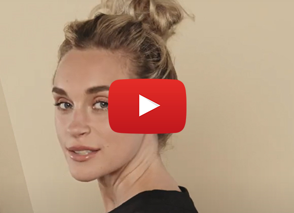 En nærbilde av en person med blondt hår oppsatt i en knute mot en beige bakgrunn, med en YouTube-knapp over bildet.