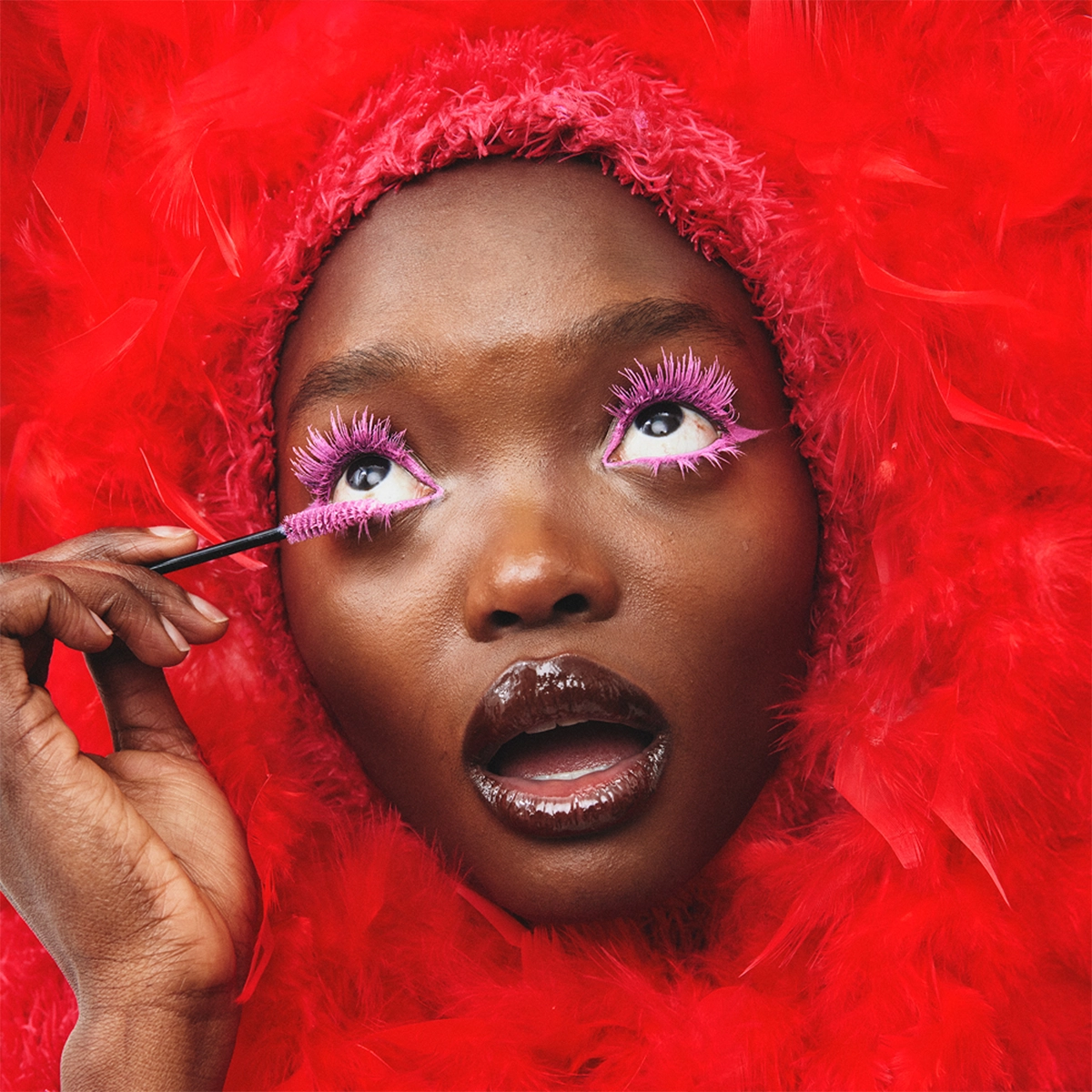 Nærbilde av en person som påfører rosa mascara på de øvre vippene med en mascarabørste. Dramatisk øyesminke med rosa vipper, glansfull lipgloss og en rød fjærhette rundt ansiktet.