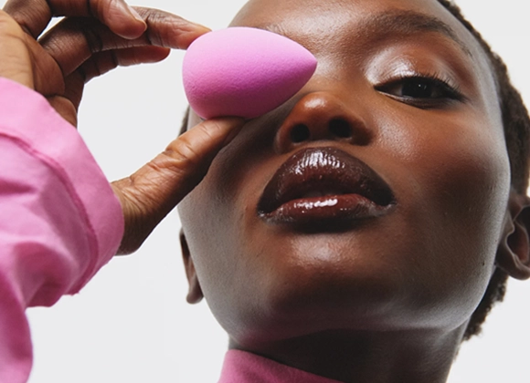 Lähikuva henkilöstä vaaleanpunaista poolopaitaa vasten vaaleaa taustaa. Henkilö pitää kädessään vaaleanpunaista meikkisientä yhden silmän edessä. Kasvot ovat hieman ylöspäin kääntyneet ja huulissa on kiiltävä meikki.