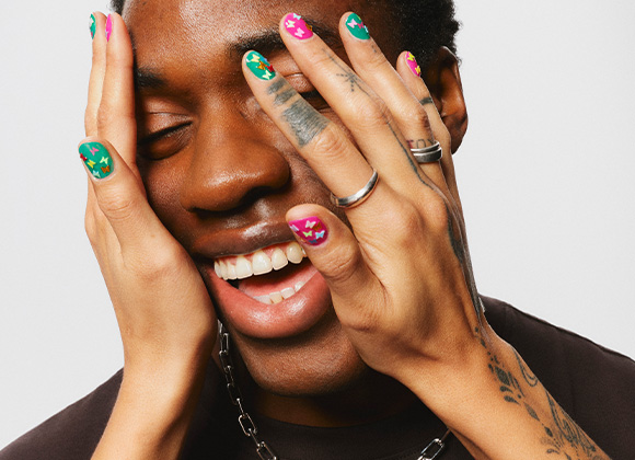  Lähikuva hymyilevästä henkilöstä, jonka kasvot peittyvät värikkäillä kynsillä koristelluilla käsillä.