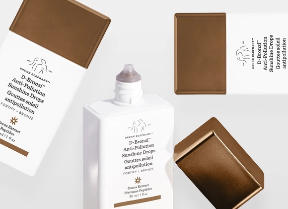 Produktbilde som viser flere flasker og korker av Drunk Elephant D-Bronzi Anti-Pollution Sunshine Drops. Hvite, firkantede flasker med bronsefargede korker i ulike posisjoner mot en lys bakgrunn.