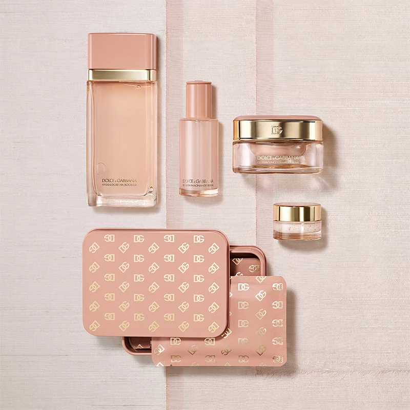 Flere kosmetikkprodukter i rosa og gull, inkludert parfymeflasker, kremer og en metallboks med Dolce & Gabbana-logoer, ligger på en lys overflate.