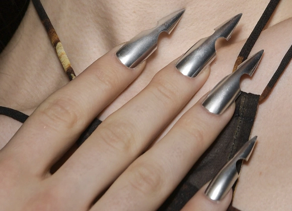 Nærbilde av en hånd med lange, metalliske negler med unik, spiss og uthult design. Neglene har en skinnende sølvfarge.