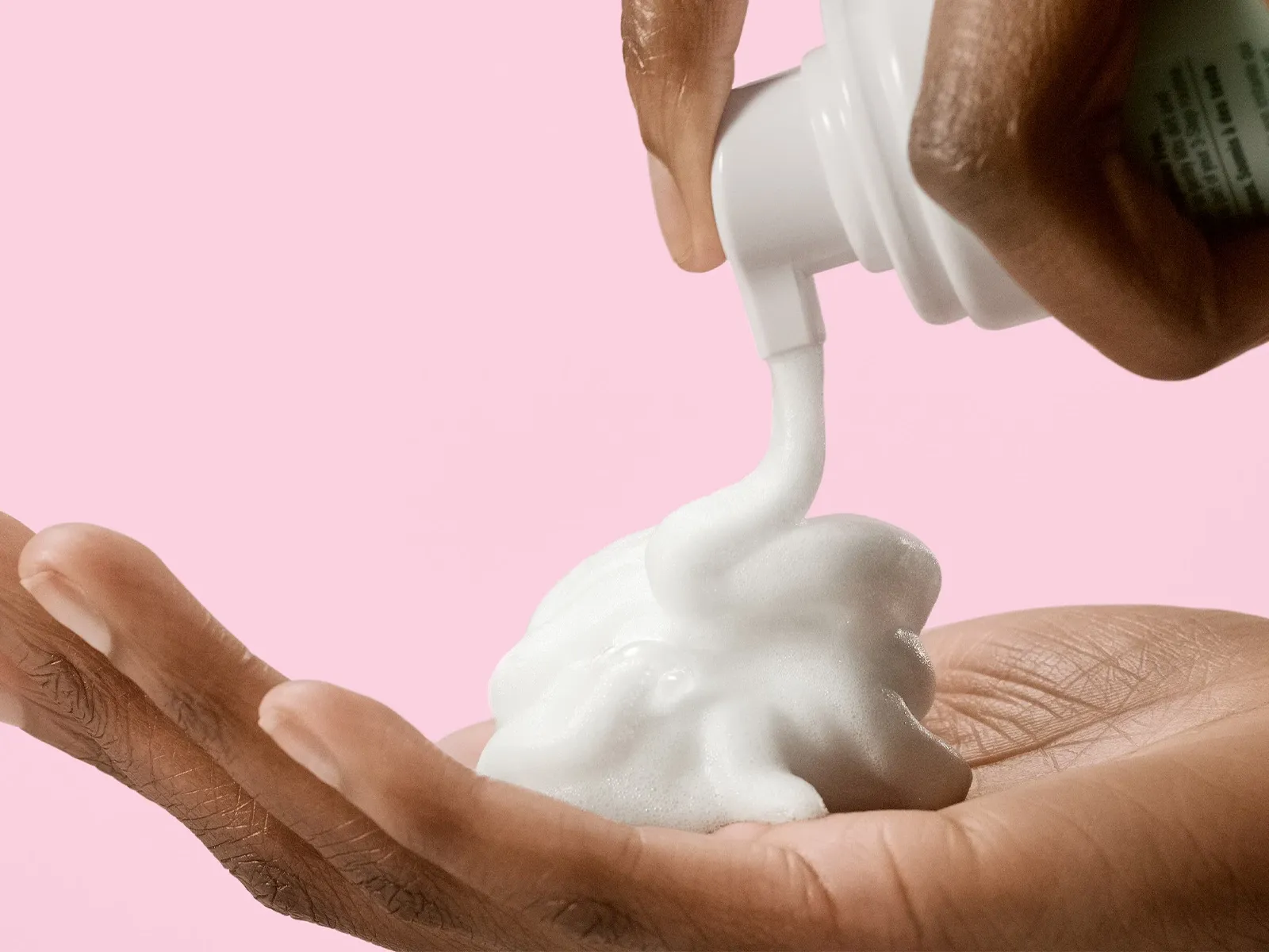 En hand som trycker ut skum från en hudvårdsprodukt till en annan hand mot en rosa bakgrund