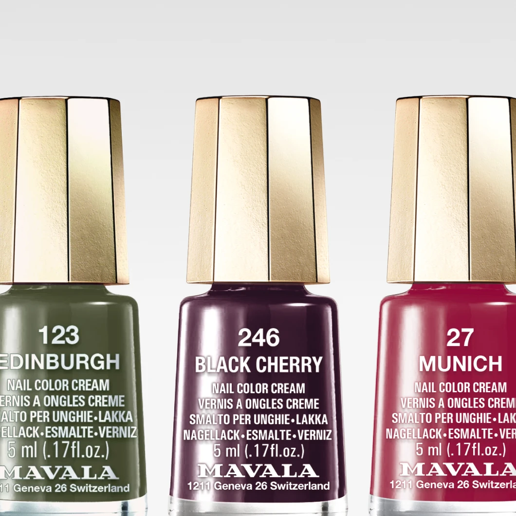 Närbild på tre Mavala-nagellack mot en ljus bakgrund: ett olivgrönt lack, ett mörkt plommonfärgat lack och ett klarrött lack står i en rak linje, alla med karakteristiska guldlock. Produkterna är centrerade och belysta för att framhäva färg och glans.
