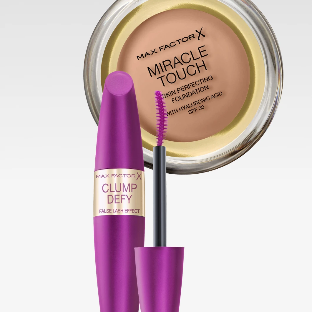 Lähikuva Max Factor -meikeistä vaaleaa taustaa vasten. Yläosassa on pyöreä Max Factor Miracle Touch Skin Perfecting Foundation -meikkivoide beige sävyssä. Edessä näkyy Max Factor Clump Defy -ripsiväri metallinhohtoisessa violetissa hylsyssä, kultainen etiketti ja esillä oleva harja kohti kameraa.