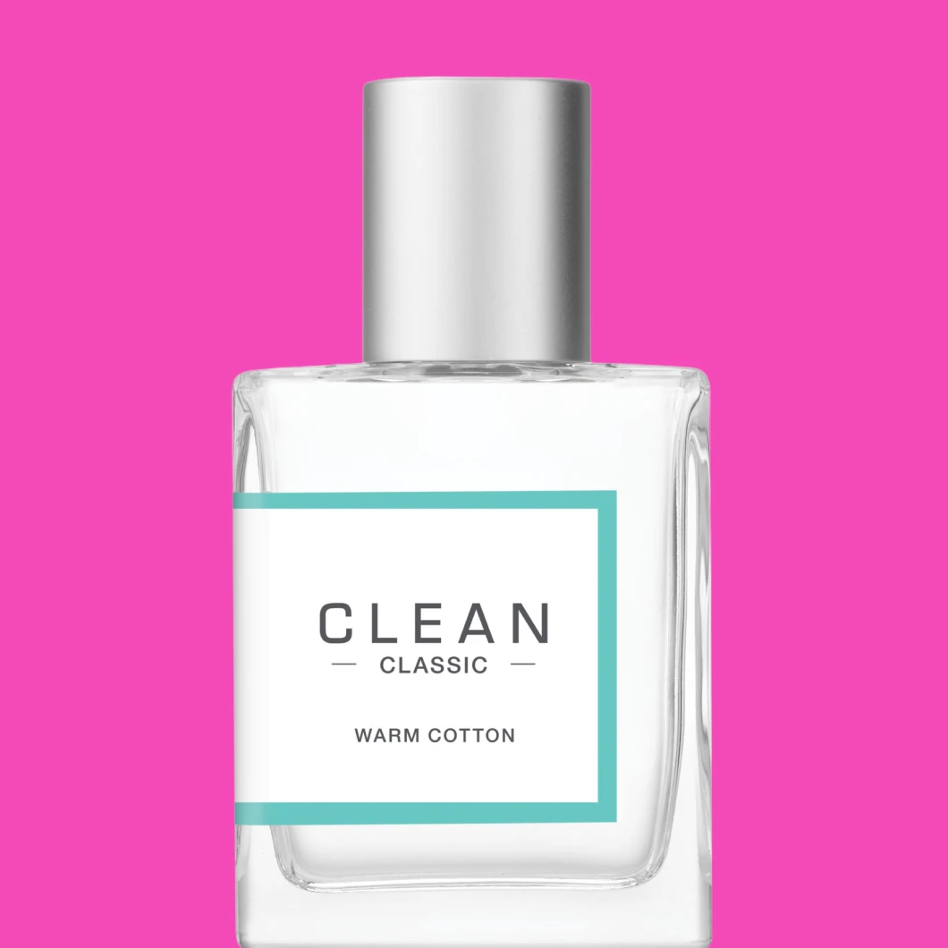 Clean Classic Warm Cotton parfymeflaske mot en sterk rosa bakgrunn, vist forfra med sølvlokk og turkis ramme rundt etiketten.