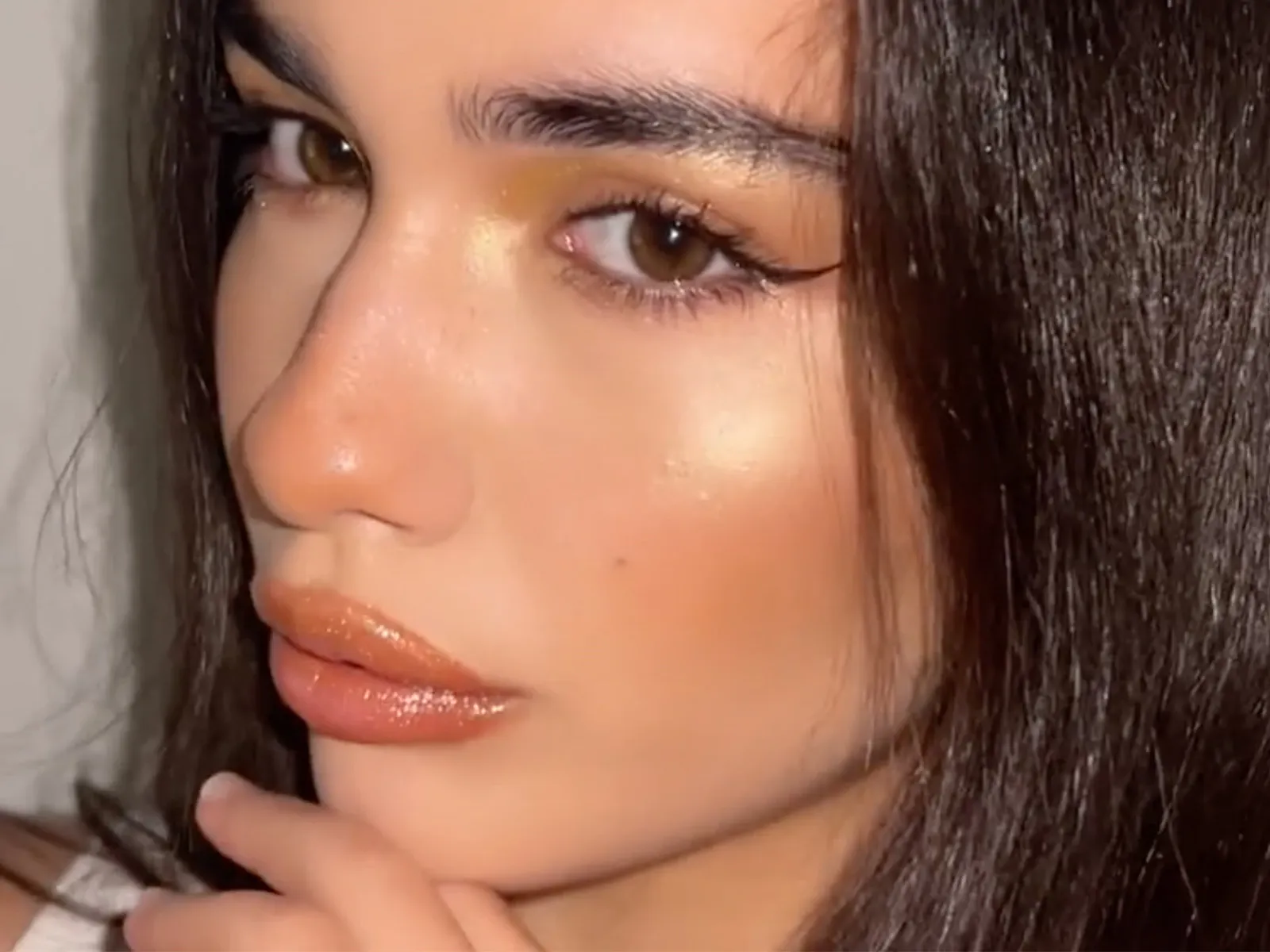 En person med långt mörkt hår håller handen vid ansiktet och har på sig en vit tröja. Hon har fylliga, rosa läppar som glänser.