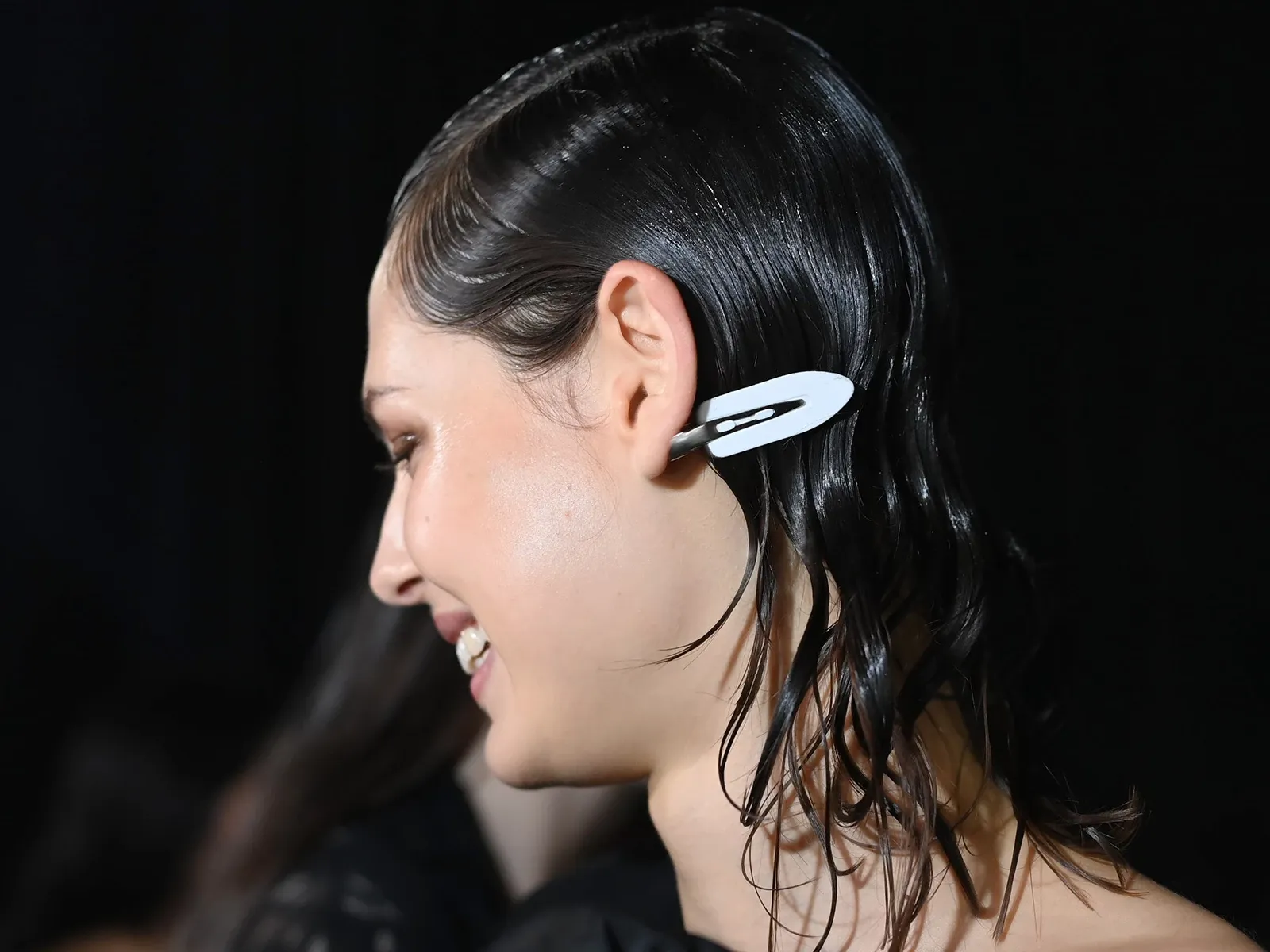 Hymyilevä henkilö, jolla on taakse kammatut märät hiukset ja hiusklipsi, tummalla taustalla.