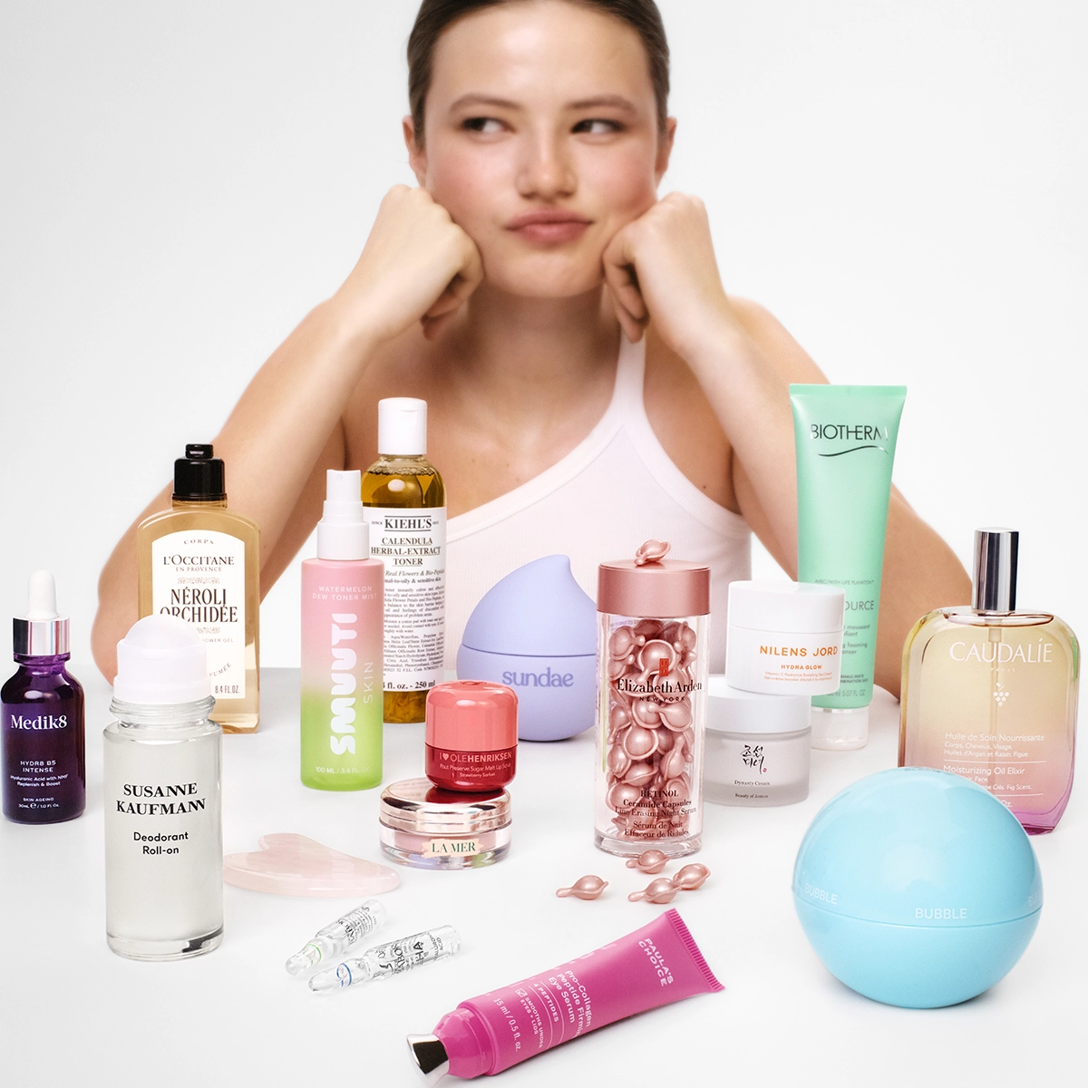 Henkilö nojaa leukaa käsiinsä pöydän takana, joka on täynnä ihonhoito- ja kauneustuotteita, kuten L’Occitane Néroli & Orchidée, Kiehl’s Calendula Herbal Extract Toner, Biotherm Aquasource, NUXE Huile Prodigieuse, Caudalie Eau des Vignes, Medik8 -seerumi, Susanne Kaufmann Deodorant Roll-On ja Elizabeth Arden -kapseliseerumi.