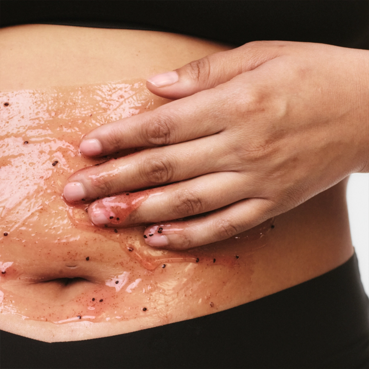 Närbild av en hand som applicerar kroppsskrubb med korn på magen, med glansig hud och synliga exfolierande partiklar inom hudvård.