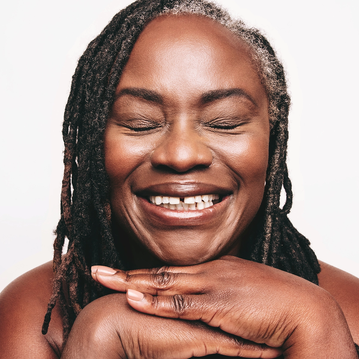 Lähikuva henkilöstä, jolla on suljetut silmät ja luonnollinen, hehkuva iho, korostaen ihonhoitoa, kuulautta ja tasaista ihon pintaa. Kädet on asetettu leuan alle korostamaan kasvojen muotoa.