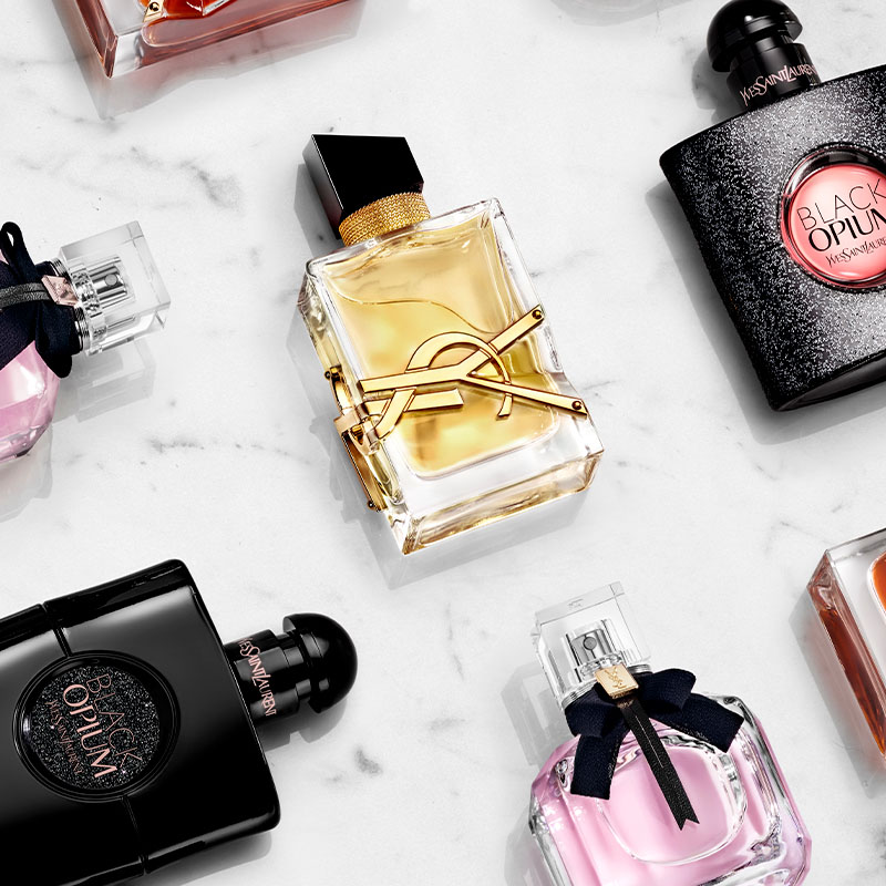 Vakre parfymeflasker fra Yves Saint Laurent. Flaskene er elegant plassert på en hvit marmorbakgrunn. Den midterste flasken er gjennomsiktig med en gullfarget væske og har YSL-logoen i gull. Rundt den står andre flasker i ulike farger og former, blant annet svart, rosa og oransje.