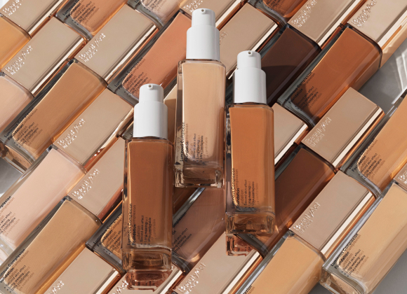 Tre foundationflasker plassert på en overflate dekket av foundations i forskjellige nyanser.