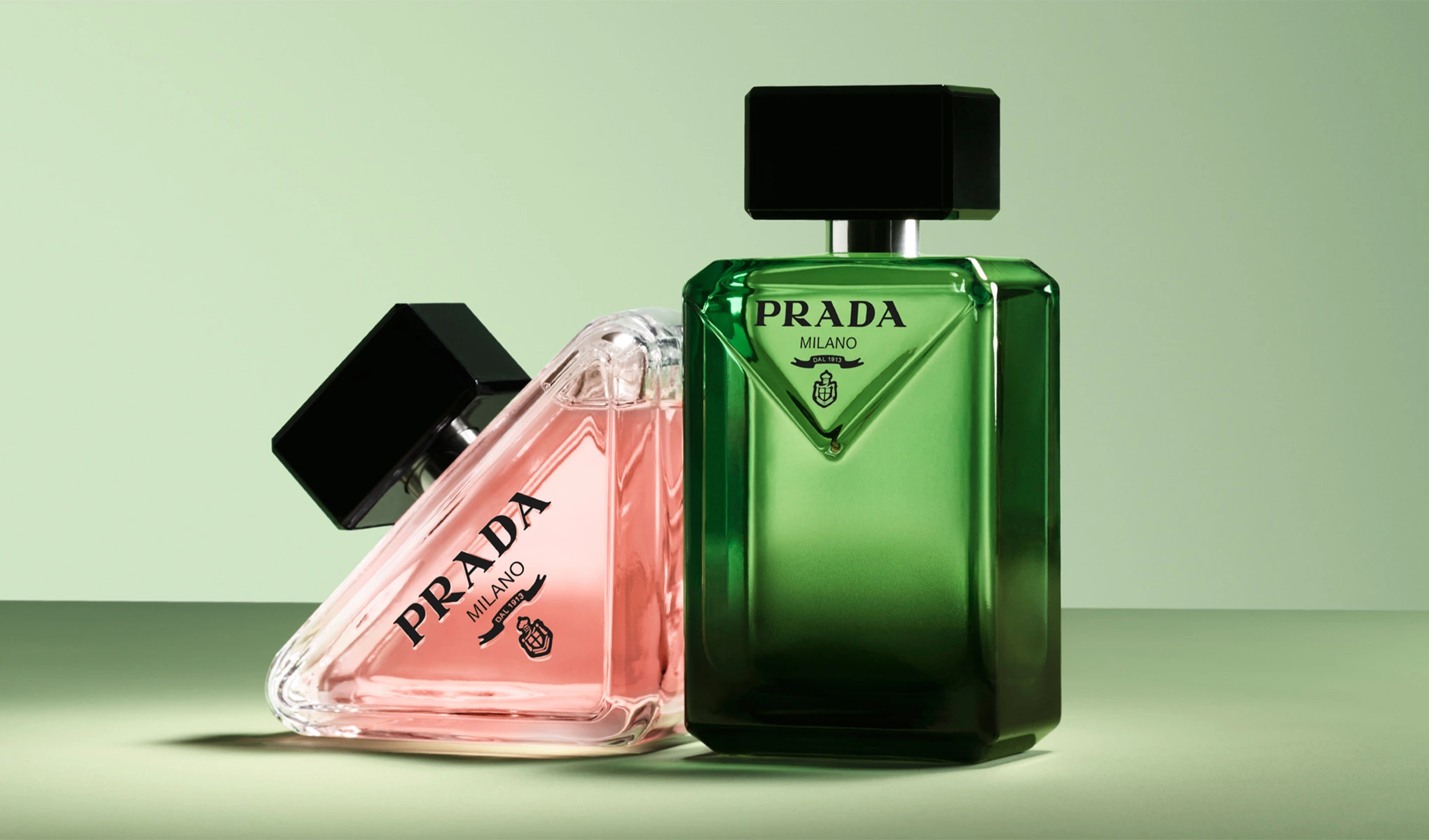 Två Prada Milano-parfymflaskor på en ljusgrön bakgrund. Till vänster en rosa flaska som ligger på sidan, och till höger en grön flaska som står upprätt. Båda flaskorna har en svart kork.