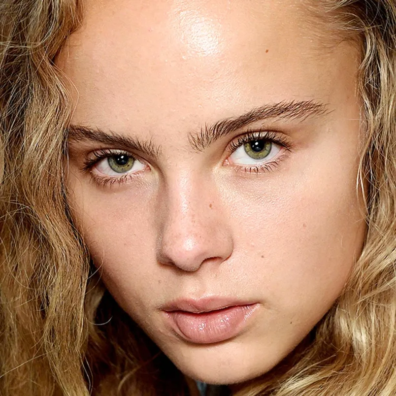 Person med vågigt blont hår och gröna ögon som tittar rakt in i kameran. Huden är klar och ser naturlig ut, med minimal eller ingen makeup.