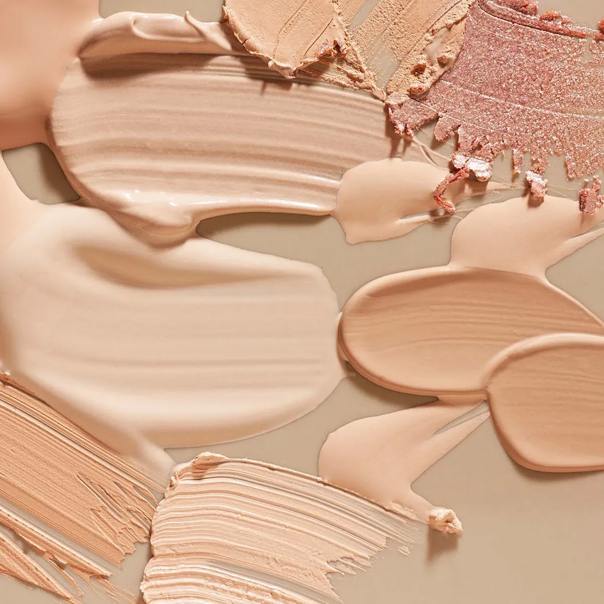 Nærbilde av forskjellige nyanser av foundation og sminke smurt ut på en beige overflate.