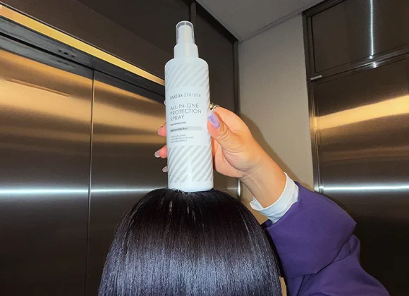 En hand håller upp Matas Striber All-In-One Protection Spray ovanpå ett glansigt, mörkt och rakt hår i en hissmiljö.