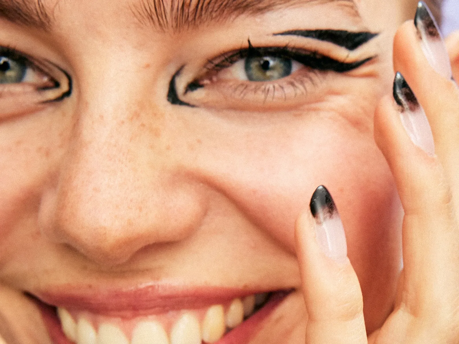 En person med svart ögonmakeup ler och håller handen vid ansiktet. Naglarna är målade med svart och vitt mönster.