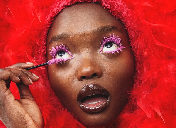 Nærbilde av en person som påfører rosa mascara på de øvre vippene med en mascarabørste. Dramatisk øyesminke med rosa vipper, glansfull lipgloss og en rød fjærhette rundt ansiktet.