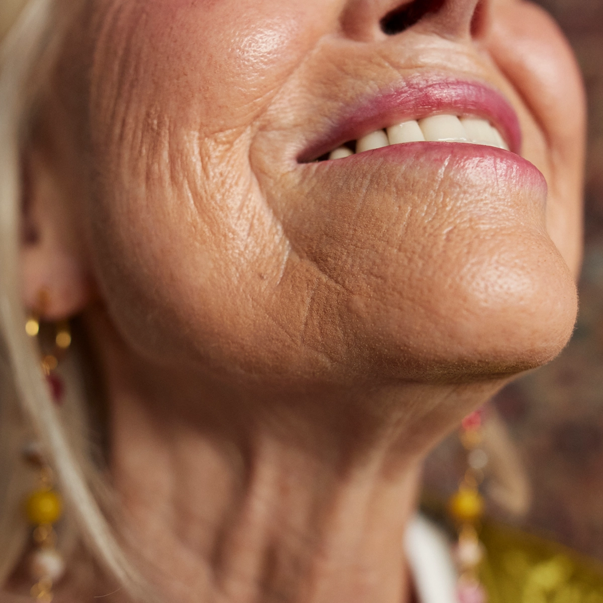 Närbild av en persons nedre ansikte och hals mot suddig bakgrund. Personen ler med lätt synliga tänder. Huden runt mun, haka och hals är tydligt synlig med naturlig struktur. Personen bär små örhängen.