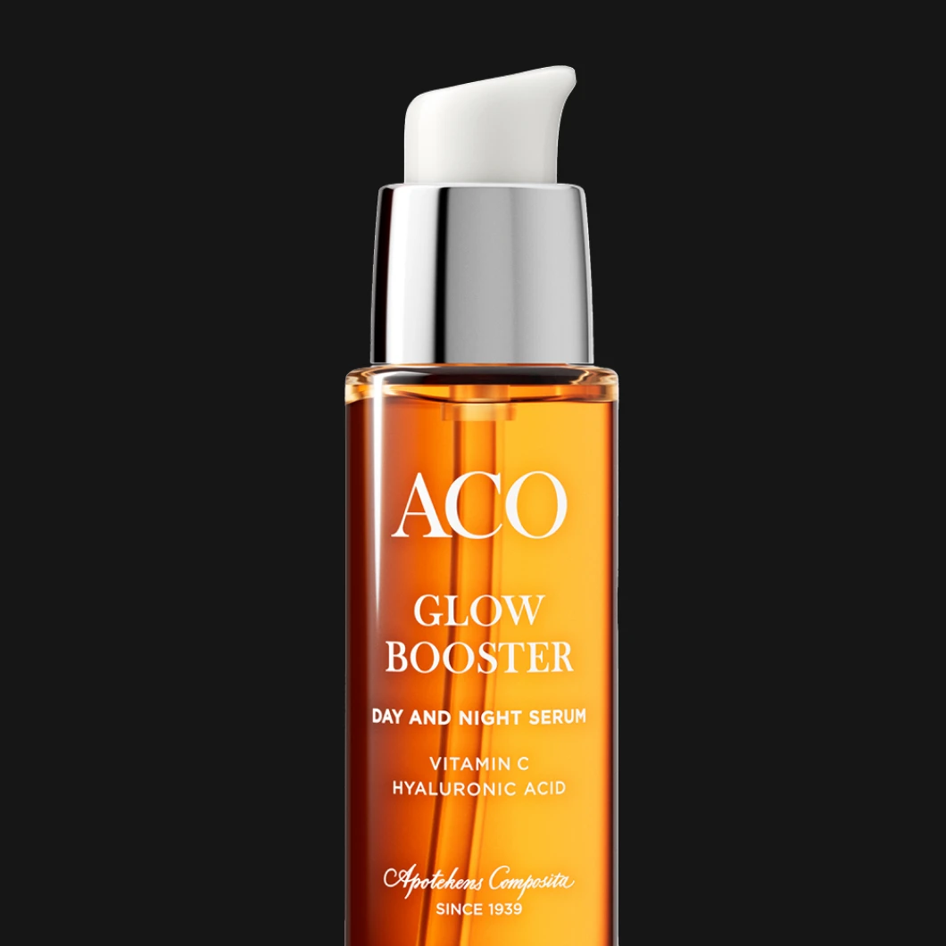 Flaska med ACO Glow Booster Day and Night Serum i genomskinlig orange förpackning med pump. Text på flaskan: “ACO Glow Booster”, “Day and Night Serum”, “Vitamin C”, “Hyaluronic Acid”. Svart bakgrund.