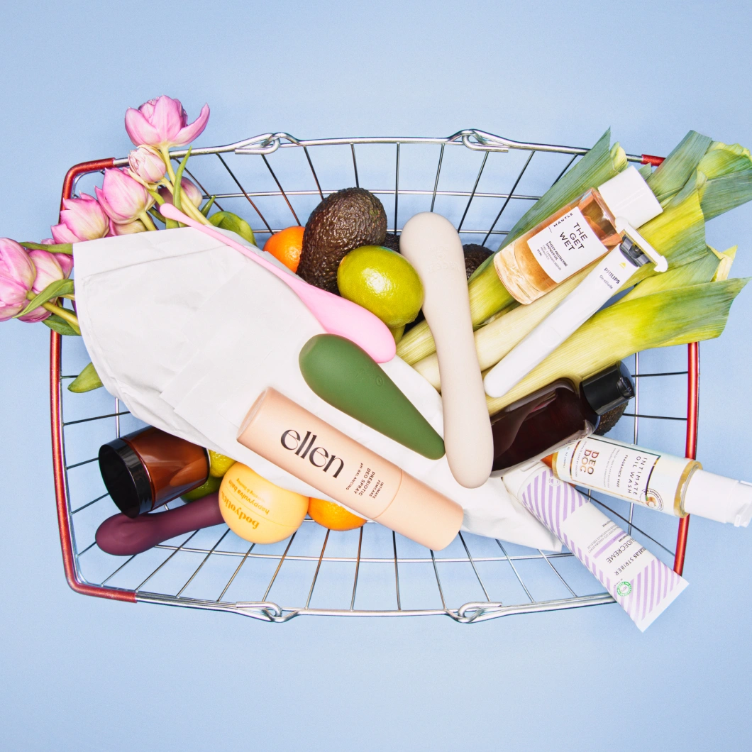 Wellness- och intimprodukter i en kundkorg tillsammans med frukt, grönsaker och blommor mot ljusblå bakgrund.