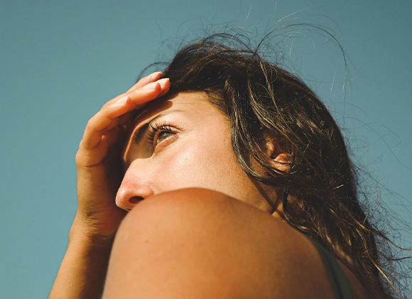 En person med langt mørkt hår holder hånden opp mot ansiktet for å skygge mot solen. 