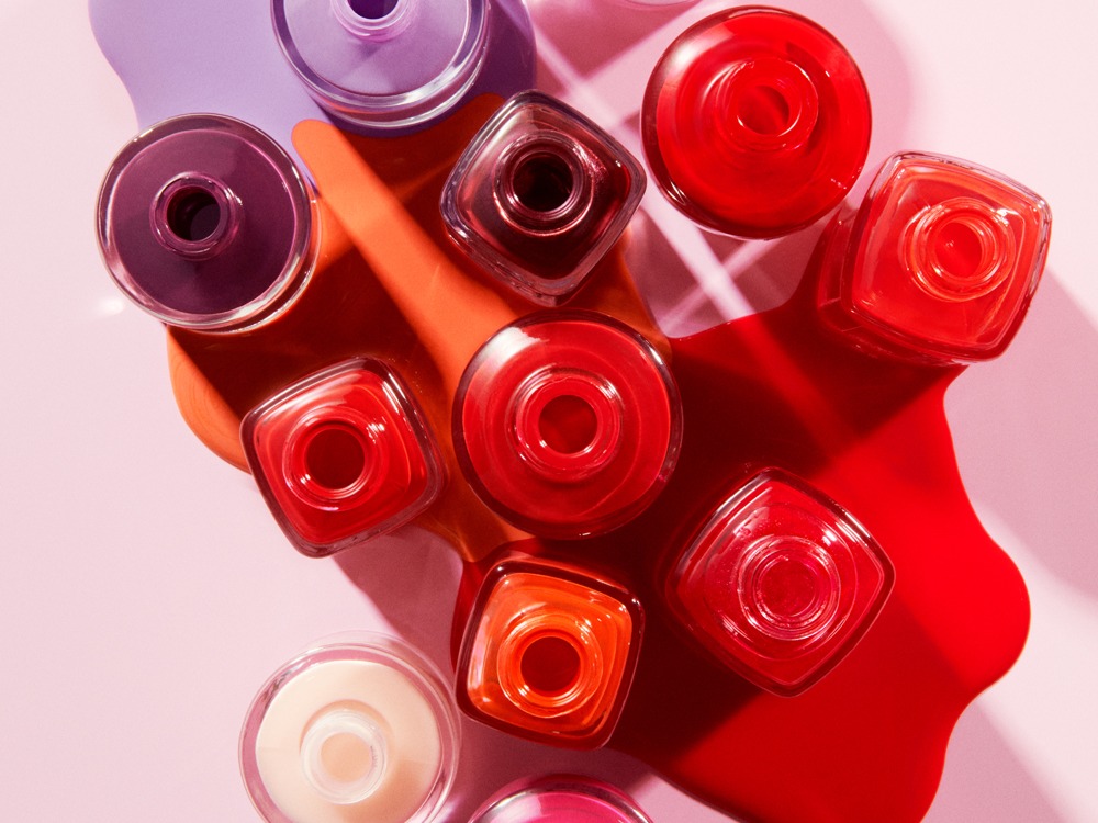Flera öppna flaskor nagellack från Atelier Rouge i olika färger, sedda ovanifrån, med färgstänk av rött, orange och lila på en rosa bakgrund.