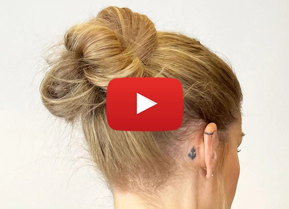 En person med blont hår uppsatt i en knut och en liten tatuering bakom örat. En stor röd YouTube-spelknapp är överlagd på bilden.