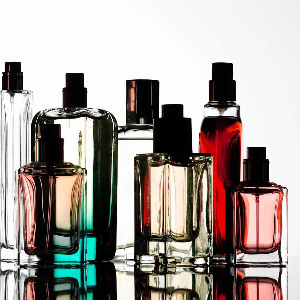 Flere parfymeflasker i ulike former og farget glass står samlet på en reflekterende overflate mot lys bakgrunn. Transparente, grønne, røde og ravfargede flasker med svarte pumper og korker gir en stilren presentasjon av parfyme og duft.
