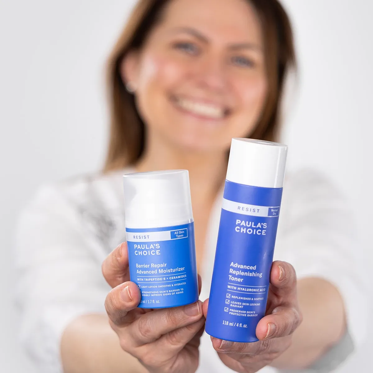 To blå produkter fra Paula's Choice foran en uklar kvinne med et stort smil mot en hvit bakgrunn.