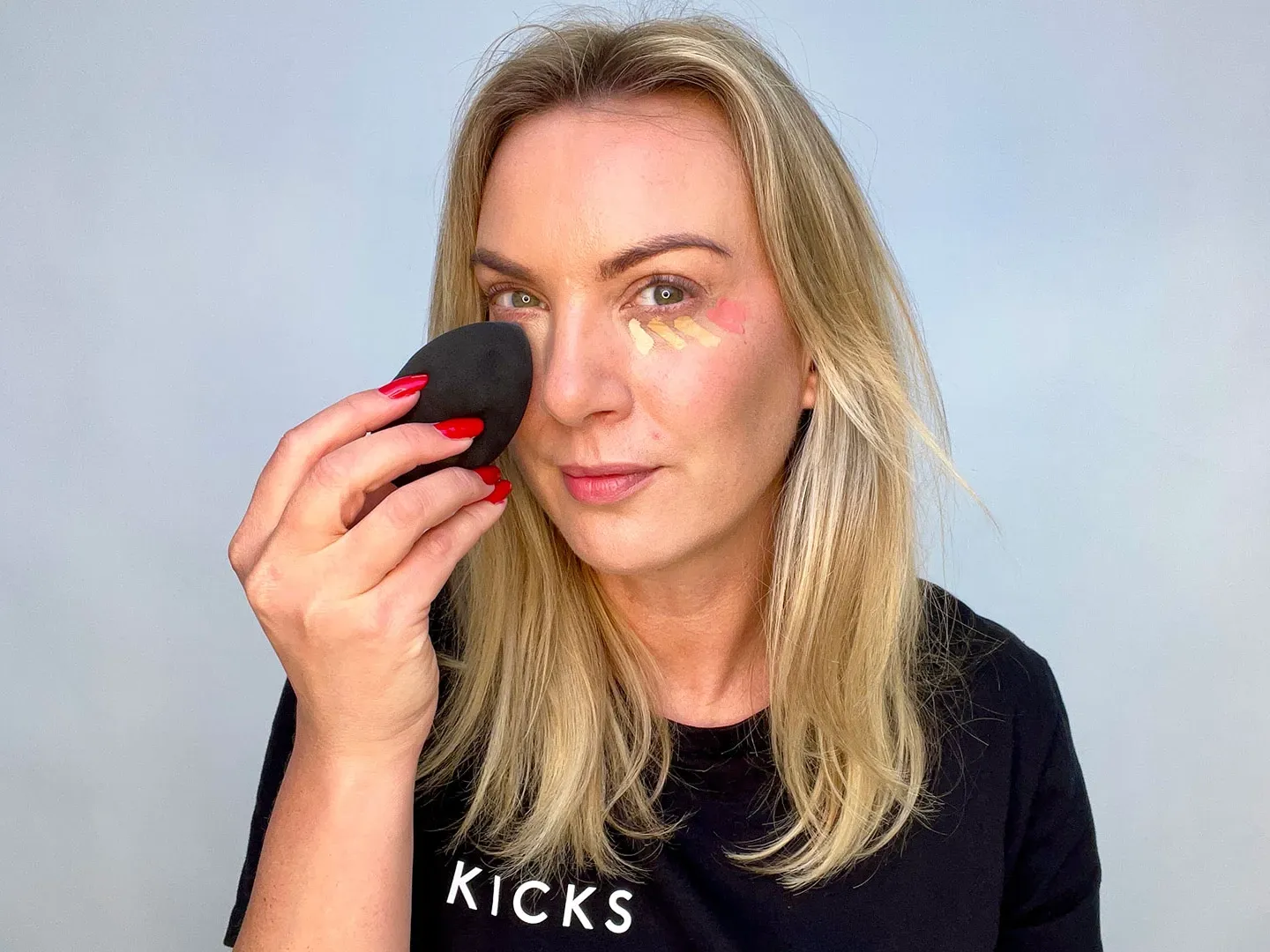 En kvinne med blondt hår holder en svart sminkesvamp i hånden. Hun har røde negler og har på seg en svart genser med teksten "KICKS".