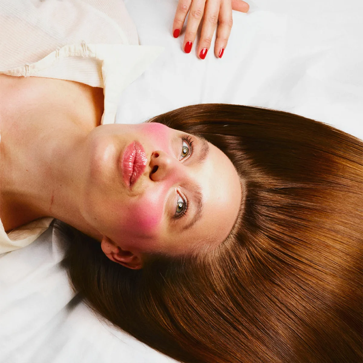En person ligger mot en hvit bakgrunn med langt, rett brunt hår spredt rundt hodet. Kinnene er markert med sterkt rosa rouge, og leppene har en blank rosafarget glans. En hånd med rødlakkerte negler vises over hodet.