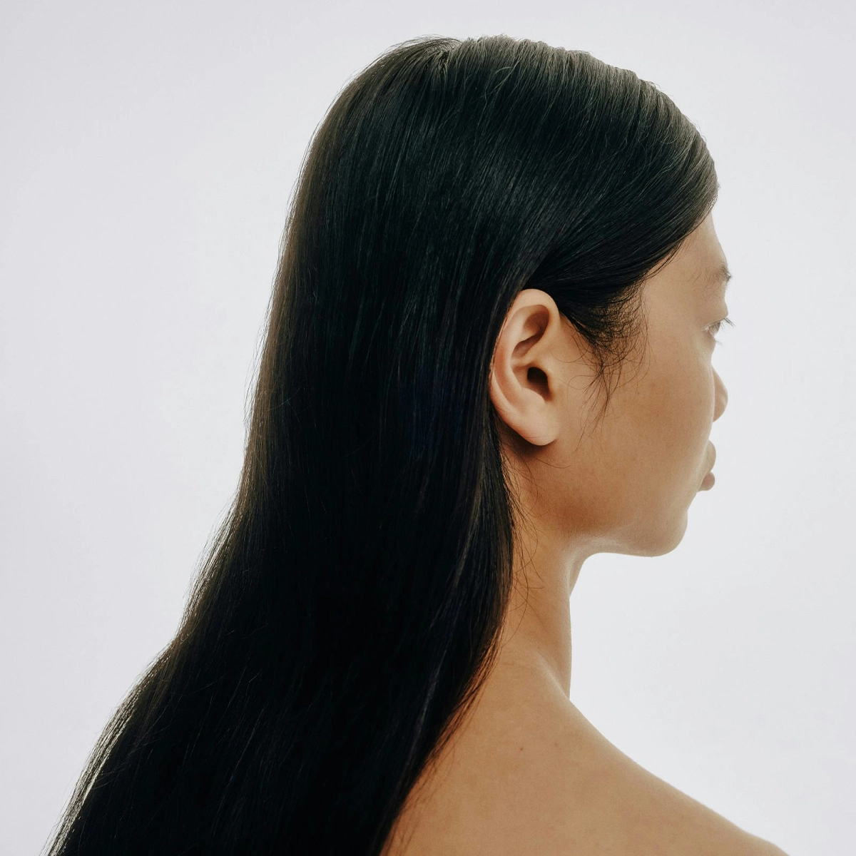 Profil av en person med langt, rett mørkt hår mot lys bakgrunn, med fokus på hårets glans og glatte struktur for hårpleie.