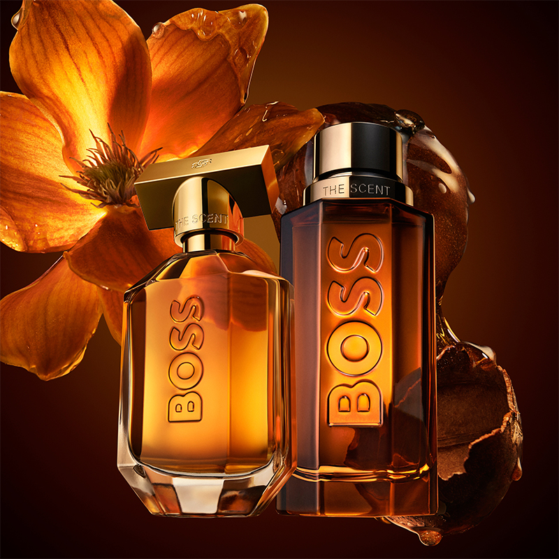 Hugo Boss The Scent-parfymeflasker i varme ravtoner mot blomsterbakgrunn.