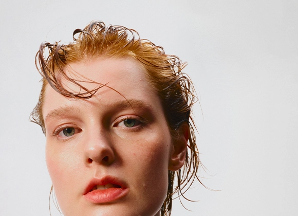 Nærbilde av ansikt med naturlig makeup og fuktig hudfinish, med fokus på hudpleie og basemakeup. Håret er kjemmet bakover og lett vått mot en lys bakgrunn.