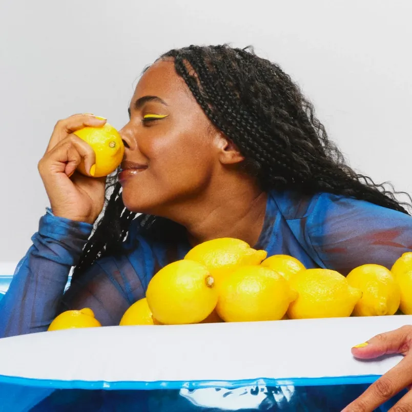 Kvinna med ljusgul eyeliner som har massor av citron framför sig. Kvinnan luktar på en citron och ler.