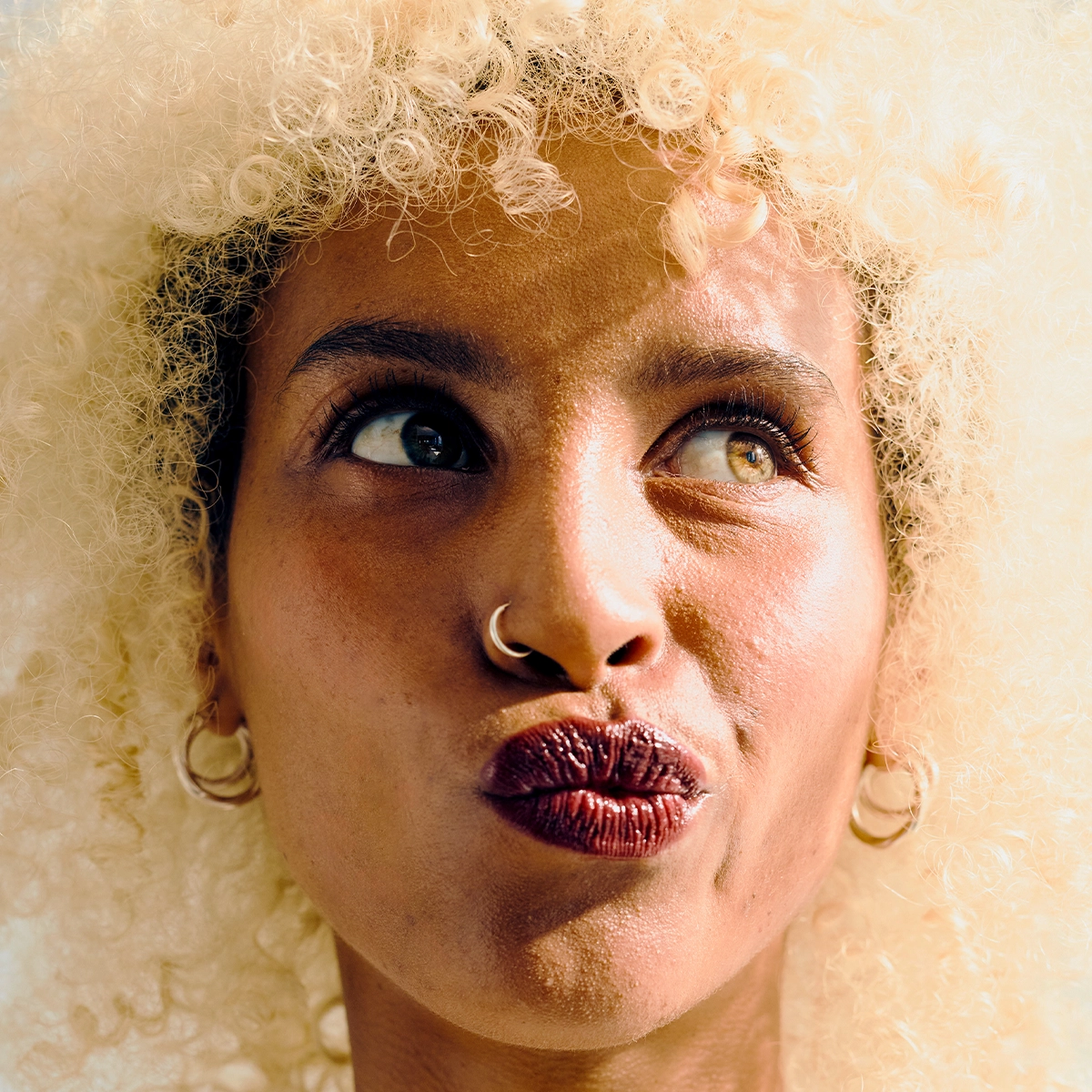 Nærbilde av en person med krøllete hår som ser opp til siden. Personen har markerte øyenbryn, mascara, mørk leppestift, nesering og øredobber mot en lys bakgrunn.