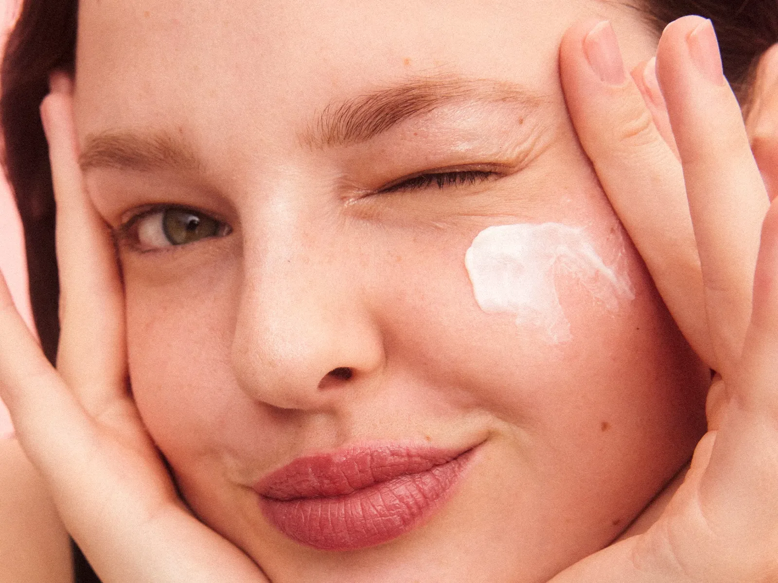 En person håller upp sina händer mot ansiktet och har en vit kräm på kinden mot en rosa bakgrund.