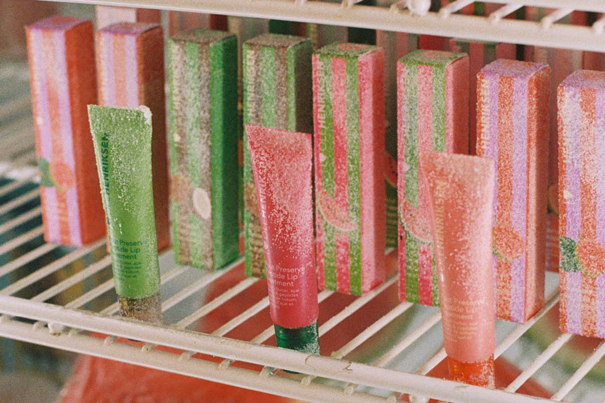 Fargerike leppeprodukter fra Ole Henriksen står på en frostet hylle sammen med matchende esker i grønt, rosa og oransje. Tubene med teksten “Ole Henriksen” og “Lip Treatment” er dekket av rim og plassert i et kjøleskapslignende miljø med vannmelon i bakgrunnen.
